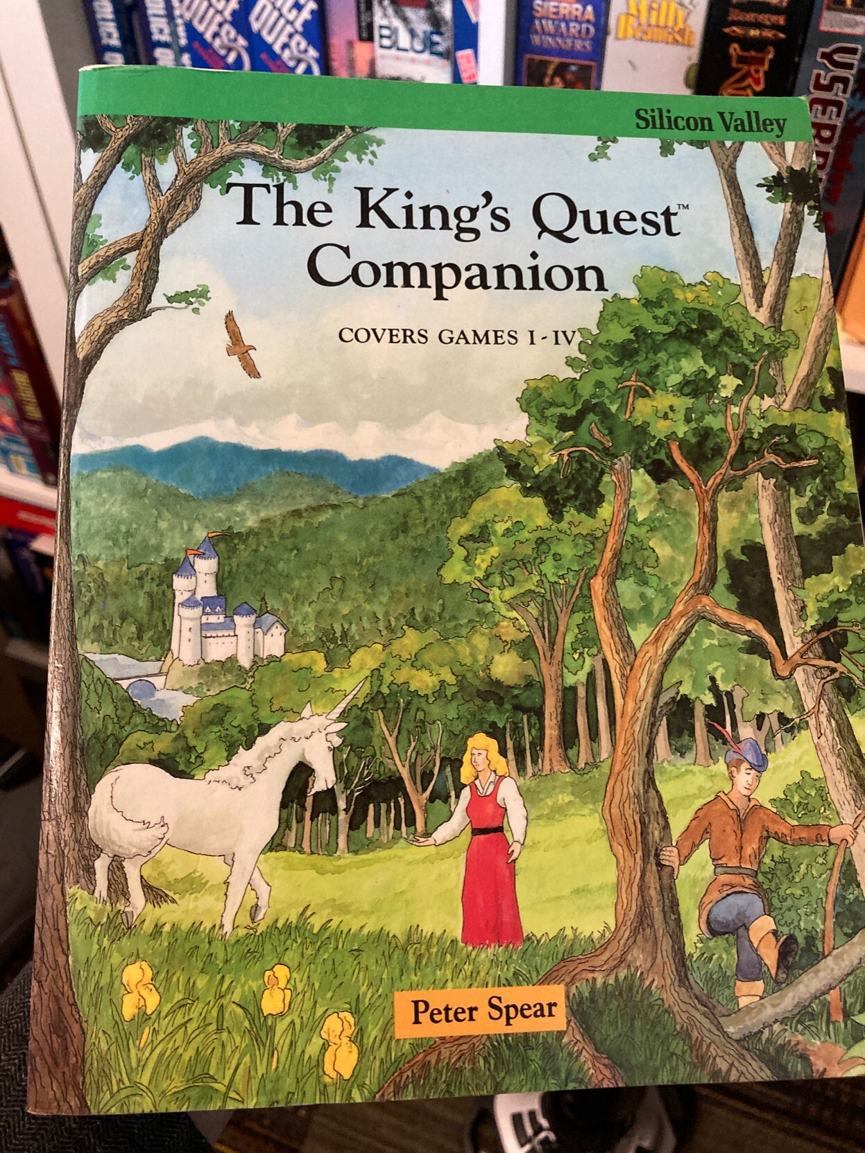 The front cover of The King's Quest Companion, by Peter Spears.

It covers games I-IV.

The art shows Princess Rosella, King Graham and a white unicorn in a verdant field, with a medieval castle in the background. The atmosphere is restful.