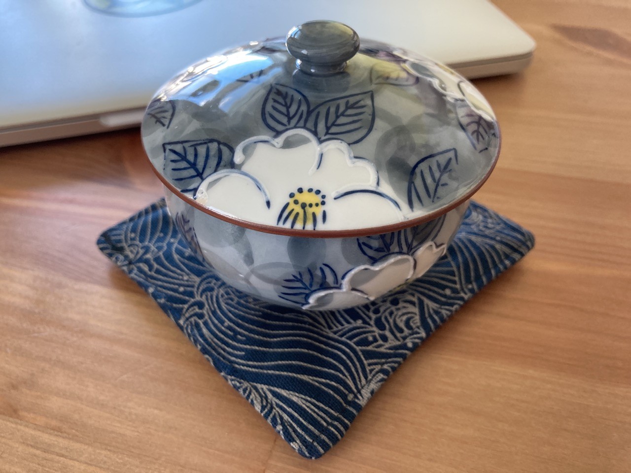 The teacup with its lid on - a white flower with yellow center. It sits upon a blue coaster with waves. 